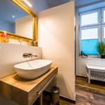 Bad mit Waschbecken auf Holzkonsole, goldgerahmtem Spiegel und Fenster mit Pflanze.