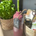 Willkommen-Schild an einer rosa Vase mit grüner Pflanze und Basilikum im Hintergrund.