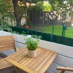 Terrasse mit Holztisch, zwei Stühlen und Pflanze. Hintergrund: grüner Zaun und Baum.