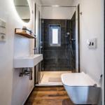 Badezimmer mit Dusche, Waschbecken und Toilette. Holzfußboden und runder Spiegel.