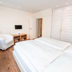 Zimmer mit Doppelbett, Kleiderschrank und Sessel. Holzboden und Wand-TV.