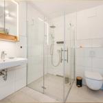 Badezimmer mit Dusche, Waschbecken und Toilette. Weiße Fliesen und Glasduschkabine.