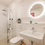 Badezimmer mit Dusche, Waschbecken und Toilette. Rundes Spiegel mit Beleuchtung.