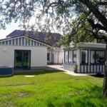 Haus mit Garten, blau-weißen Fenstern und großen Fenstern. Großer Baum im Vordergrund.
