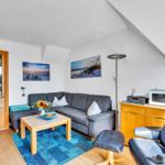 Wohnzimmer mit Sofa, Sessel und Küchenschränkchen. Blauer Teppich und Wandbilder.