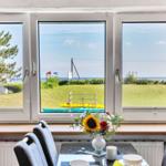 Grosses Fenster mit Blick auf Grasland und Strand. Tisch mit Stühlen und Blumen.