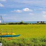 Kajaks stehen auf einer grünen Wiese vor einem Strand mit Meer.