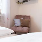 Ein Bett mit weißer Bettwäsche, ein brauner Sessel mit Kissen und ein Fenster mit weißen Vorhängen.