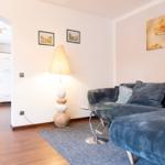 Wohnzimmer mit Sofa, Stehlampe und Glas-Tisch. Küche im Hintergrund sichtbar.