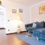 Wohnzimmer mit Sofa, Beistelltisch und Stehlampe. Küche im Hintergrund sichtbar.