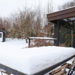 Schnee bedeckt einen Tisch und Stühle vor einem Haus mit schwarzem Dach und einem Garten.