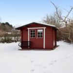 Red wooden house with snow-covered ground and a black chair in front of the door.