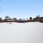 Snow-covered landscape with a red house and smaller buildings in the background.