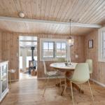 Dining room with wooden walls, round table, chairs, windows, and view into living area.