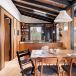 Dining area with wooden table and chairs, kitchen in background, wood walls and floor.