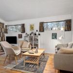Wohnzimmer mit Sofa, Sesseln, Couchtisch und Bücherregal. Holzboden und Fenster mit Vorhängen.