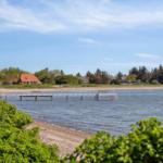 Hafen mit Steg, Strand und Häusern im Hintergrund unter blauem Himmel.