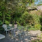 Terrasse mit Tisch und Stühlen, umgeben von grüner Vegetation und einem Haus mit Schilfdach.