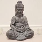 Eine Buddha-Statue sitzt auf dem Boden, flankiert von zwei Gläsern mit Kerzen.