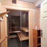 Sauna mit Holzwand, Glaswand und Holzbänken. Nebenraum mit Holzregal und Tür.