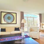 Wohnzimmer mit Sofa, Sessel und großem Fenster. Wandbild und antiker Schrank sind sichtbar.