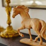 Holzfigur eines laufenden Pferdes auf dunklem Möbelstück mit goldenen Dekorationsobjekten im Hintergrund.