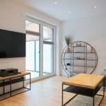 Wohnzimmer mit Fernseher, Tisch und Schrank. Große Fensterfront.