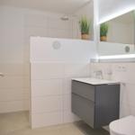 Badezimmer mit Dusche, Waschbecken und Toilette. Helle Fliesen und Spiegel mit Beleuchtung.