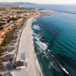 Strand mit Sand und türkisfarbenem Meer, Küstenstadt mit Häusern und Hafen im Hintergrund.