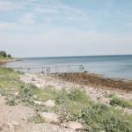 A pier leads to the sea. Shore with stones and grass. Sky with clouds.