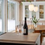 A table with wine bottle and glasses in dining area with wooden wall and window.