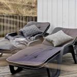 Zwei Liegestühle mit Kissen und Decke auf einer Holterrasse neben einer weißen Wand.