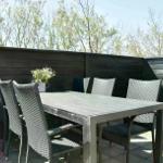Terrasse mit Tisch und sechs Stühlen, umgeben von dunklem Holzzaun und Bäumen.