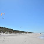Strand mit Sanddünen und Gleitschirmfliegern am Horizont.