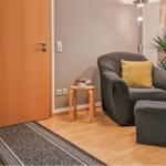 Ein Wohnbereich mit Sessel, Beistelltisch und Pflanzen. Holzboden und Tür sind sichtbar.