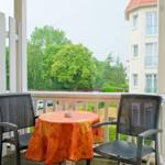 Balcony with table and two chairs, covered with orange tablecloth, overlooking green trees and building.