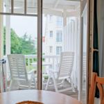 Durch Glastüren führt ein Balkon mit weißen Stühlen und Tisch. Außen sind Bäume und ein Gebäude sichtbar.