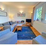 Wohnzimmer mit Sofa, Sesseln und blauem Teppich. Fenster mit Blick auf Grün.