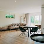 Wohn- und Essbereich mit Sofa, Tisch und Stühlen. Holzboden und große Fenster.