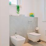 Badezimmer mit Toilette, Bidet und Waschbecken. Fenster mit Jalousie und dekorativen Gegenständen.