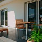 Terrasse mit Tisch, Stühlen und Liege. Glasfront und Pflanzen sind sichtbar.