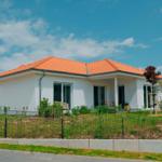 Modernes Haus mit orangefarbenem Dach und weißer Fassade. Gartenbereich mit Zaun und Sitzgelegenheit.
