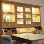 Holz-Speisekammer mit Glasfronten, Gläsern und grünen Flaschen. Holztisch und Stühle.