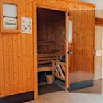 Sauna mit Holzbank und Eingangstür. An der Wand sind Steuerungseinheit und Anleitung angebracht.