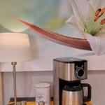 Kaffeemaschine mit Zubehör auf gelbem Tisch neben Wandbild mit Lilie.