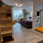 Schlafzimmer mit Bett, Fenster und Spiegel. Regal und Sitzgelegenheit sichtbar.