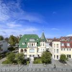 Haus mit grünem Dach und Turm in städtischer Umgebung unter blauem Himmel.