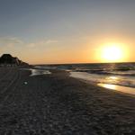Sonnenuntergang am Strand mit Wellen und Sandstrand. Gebäude sind im Hintergrund sichtbar.