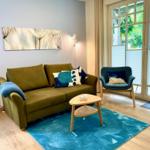 Ein Wohnzimmer mit grünem Sofa, blauem Teppich und Sitzgelegenheit vor Fenster.
