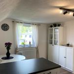 Esszimmer mit Tisch, Schrank und Fenster. Decke geneigt, Wanduhr und Pflanzen.